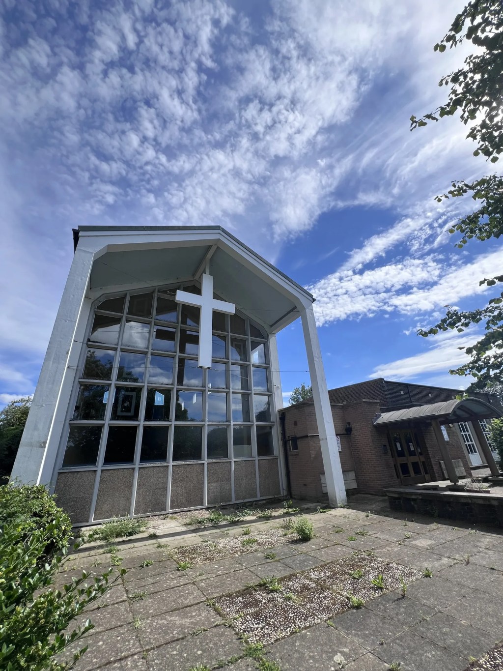 A low-angle view of a mid-century modern church facade features a towering grid of window panes beneath a stark white cross. Wispy clouds streak across a bright blue sky above the quiet, stone-paved forecourt.