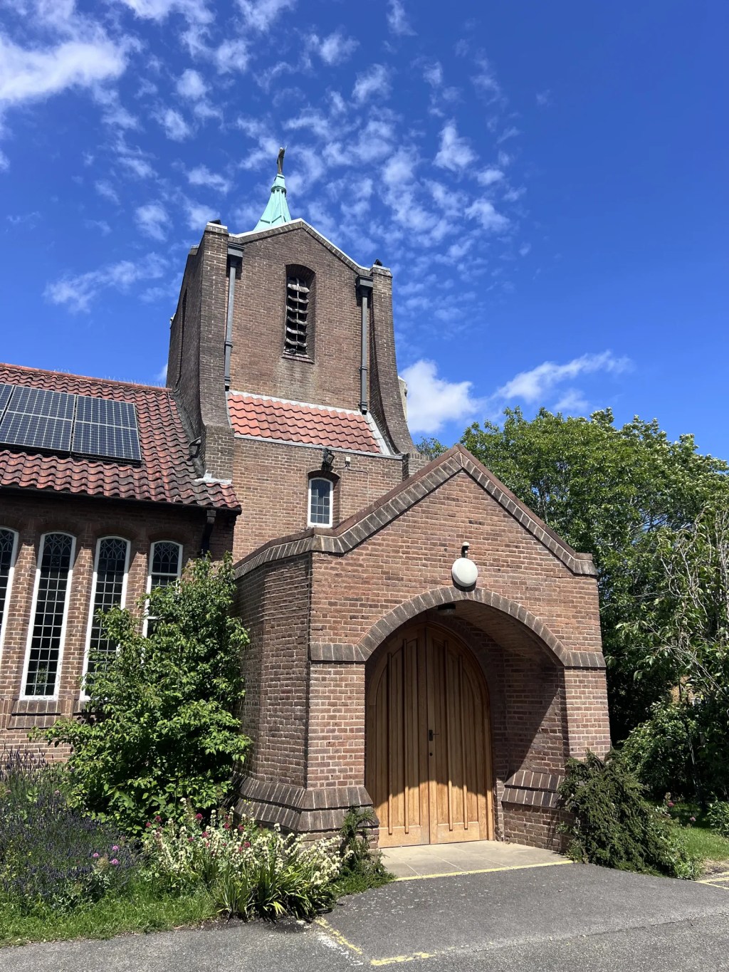 A 20th-century brick church features a gabled entrance porch with arched wooden double doors. A square brick tower rises above, capped by a small green copper spire, while solar panels sit on the red-tiled roof.