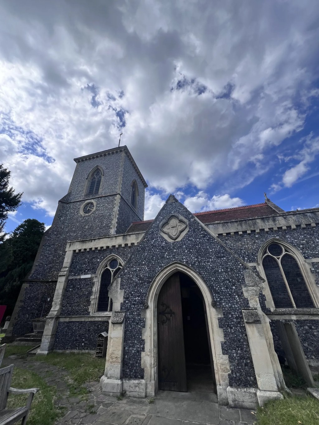 An old stone church with a prominent square bell tower rises under a dramatic, cloud-filled blue sky. The arched wooden entrance door sits slightly ajar, welcoming onlookers into the historic flint-walled building.